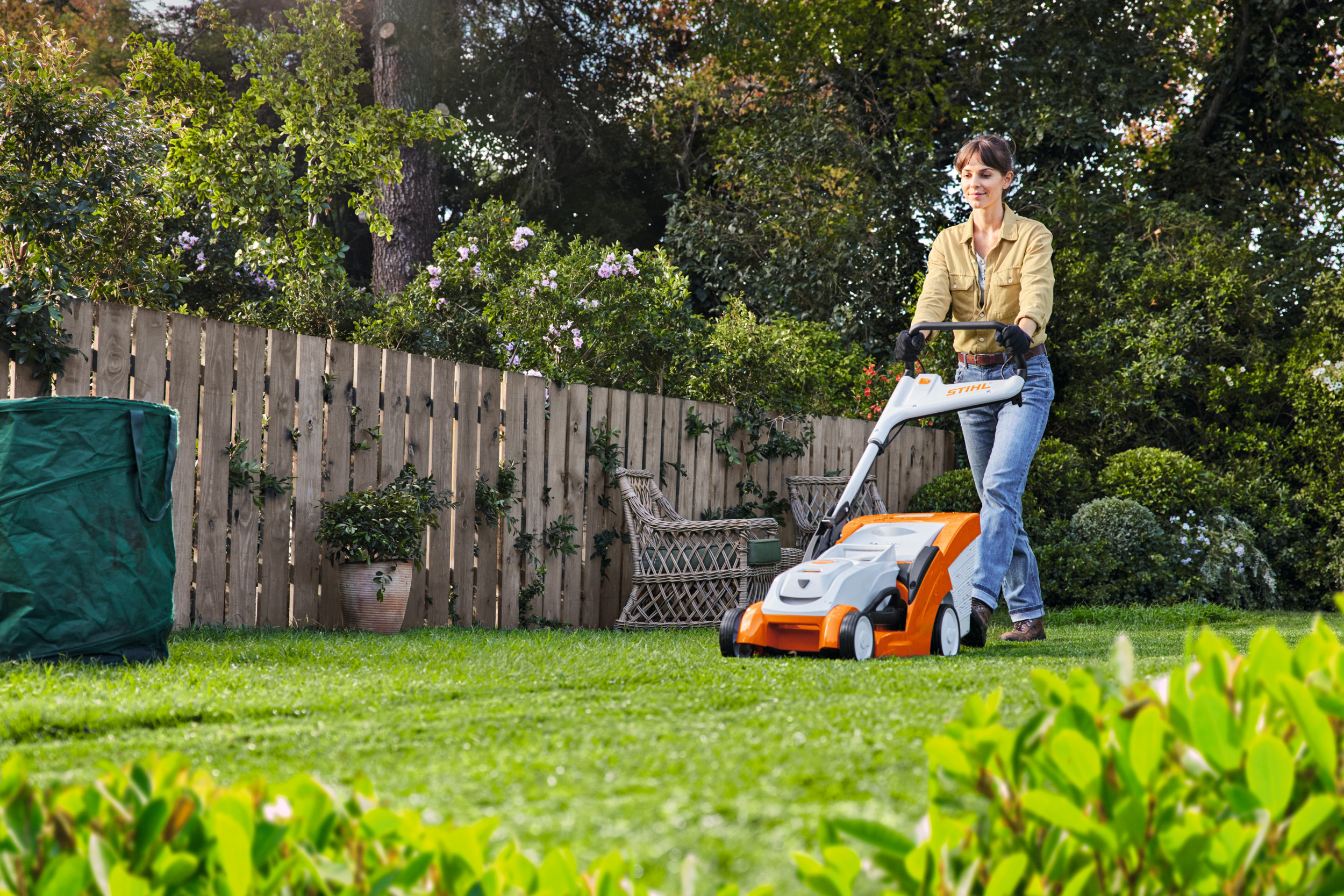 A woman is halfway through mowing her lawn using a STIHL RMA 239 C cordless lawn mower, in a garden surrounded by tall shrubs and plants