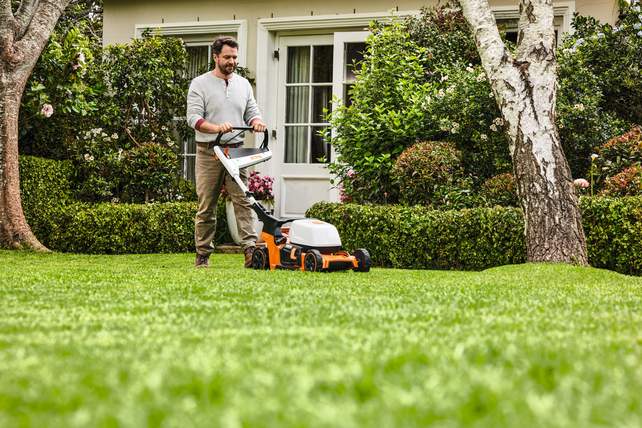 A man using a STIHL  petrol mulching mower on a lawn bordered with bushes and grasses